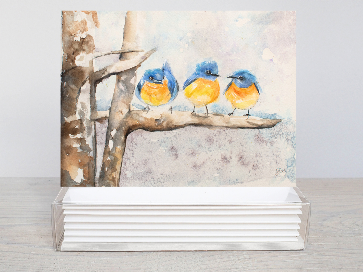 A watercolor art print featuring three bluebirds perched on a tree branch, displayed upright in a clear acrylic stand on a light wood surface against a neutral gray background.