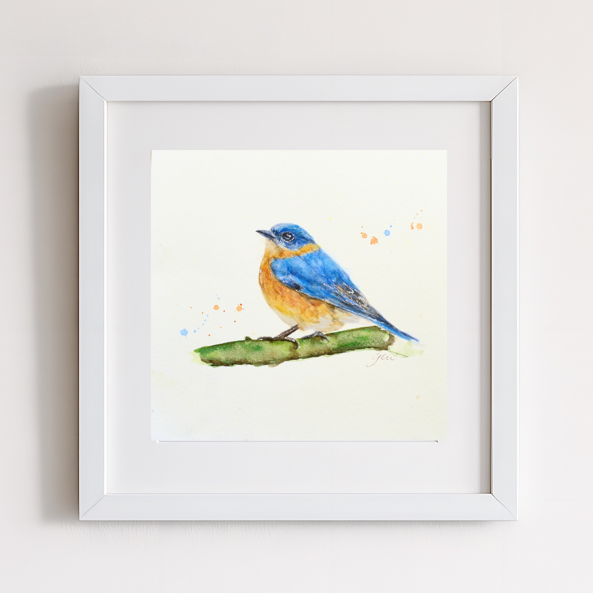 A framed watercolor painting of a bluebird perched on a green branch, set in a white frame against a light gray wall.